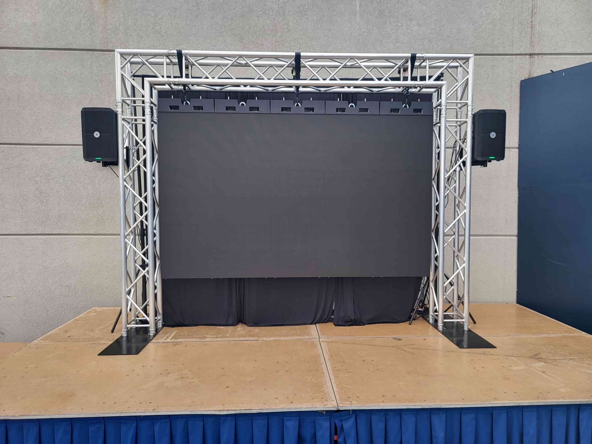 Stage with a large LED screen, silver truss frame, speakers, and a blue draped base.