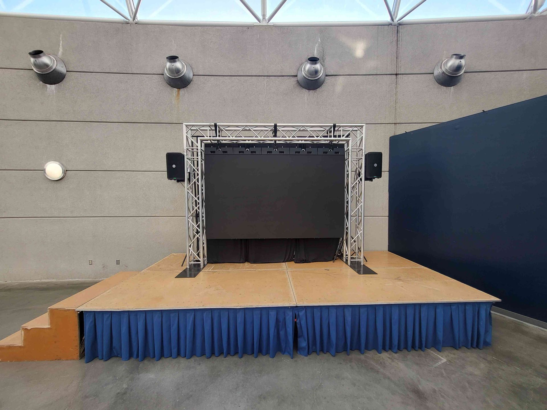 Stage with a black screen and sound equipment, in front of a blue curtain.