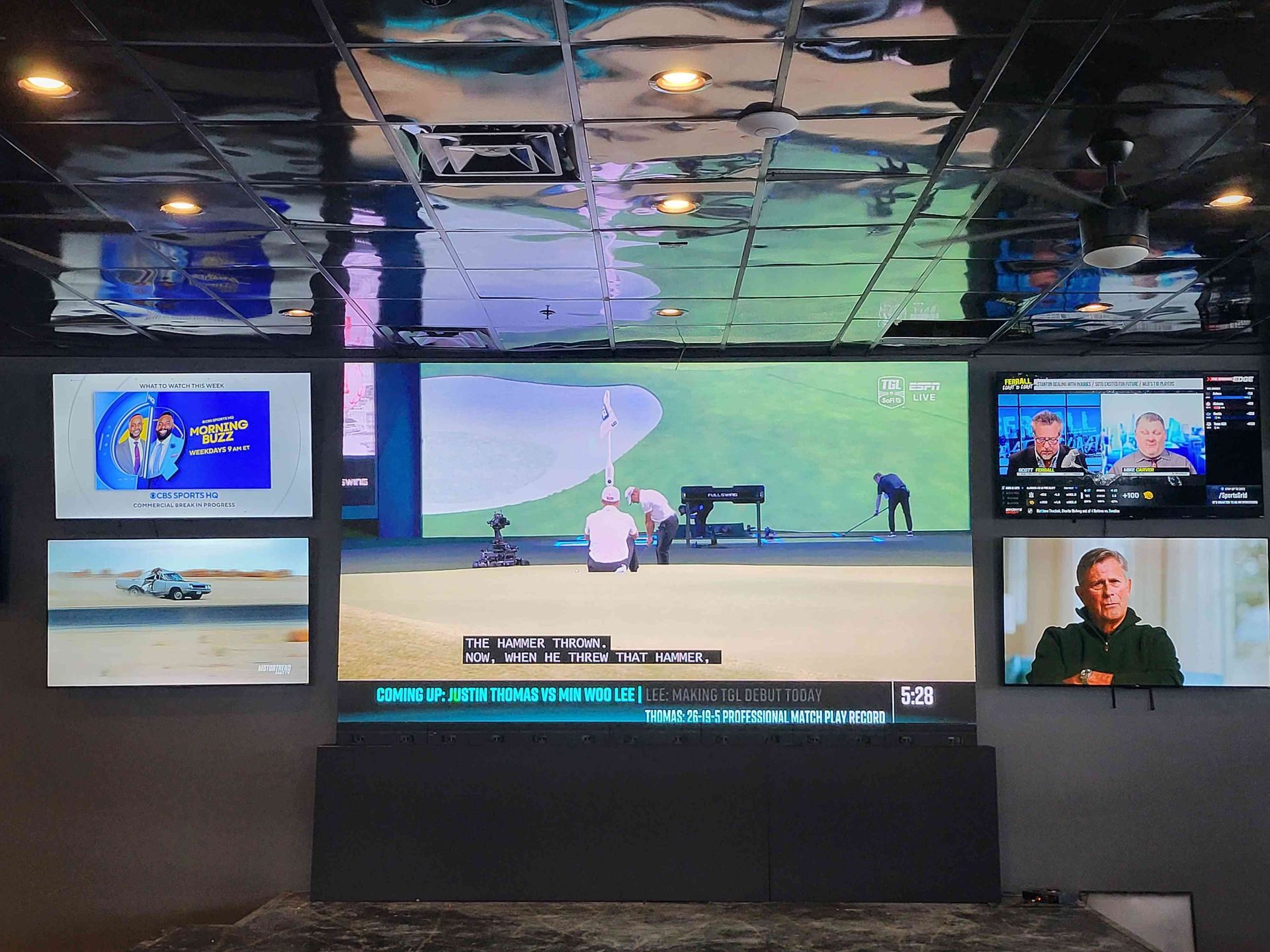 Multiple TVs showing golf and news reports on a gray wall. A reflective ceiling is above.