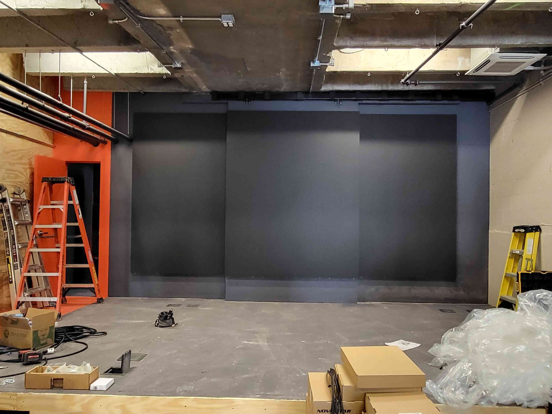 Interior view of a room under construction with dark gray wall, orange door frame, ladder, and boxes on floor.