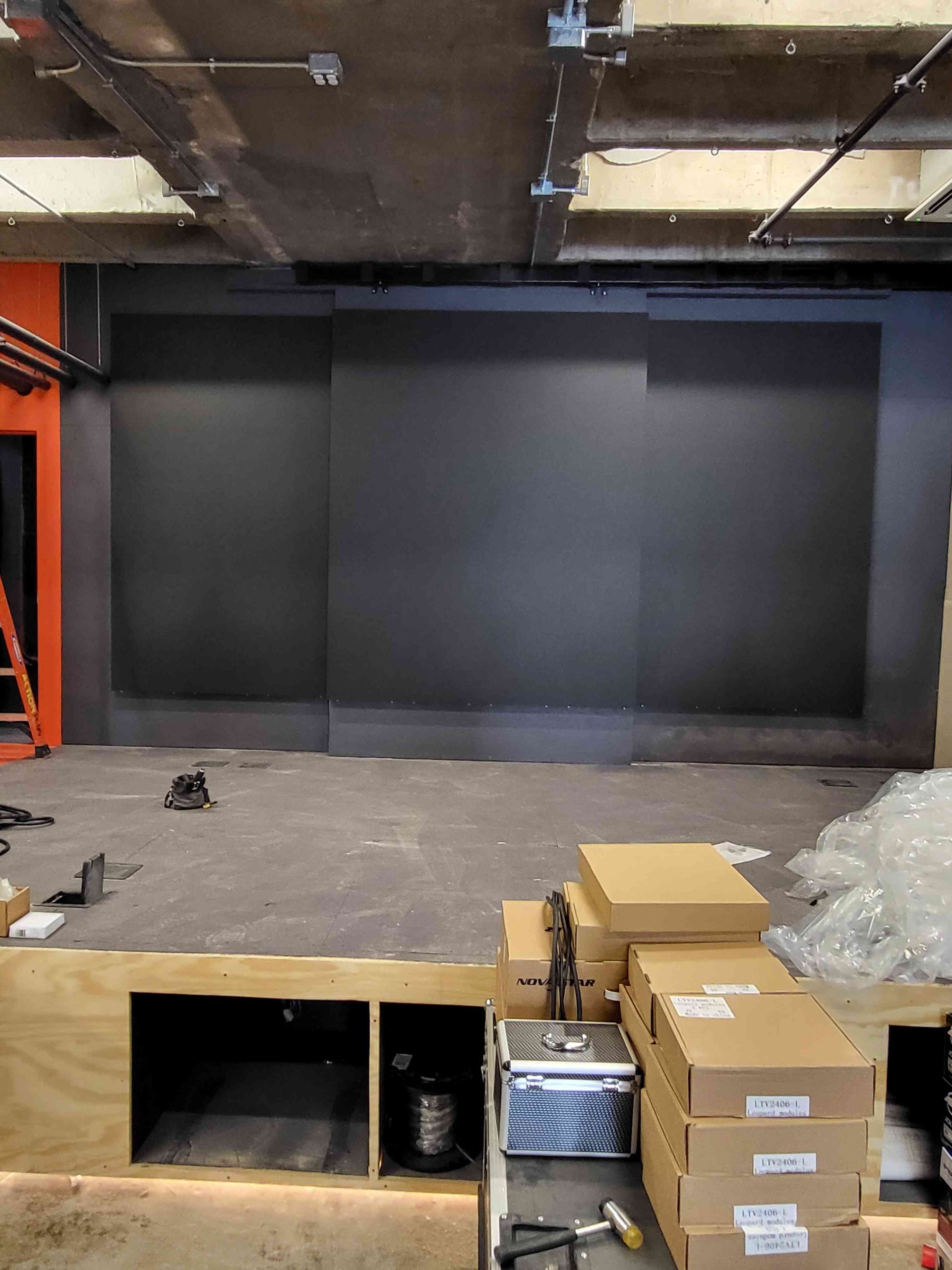 Interior of a commercial space under construction; dark gray wall, exposed ceiling, boxes, and a wooden platform.