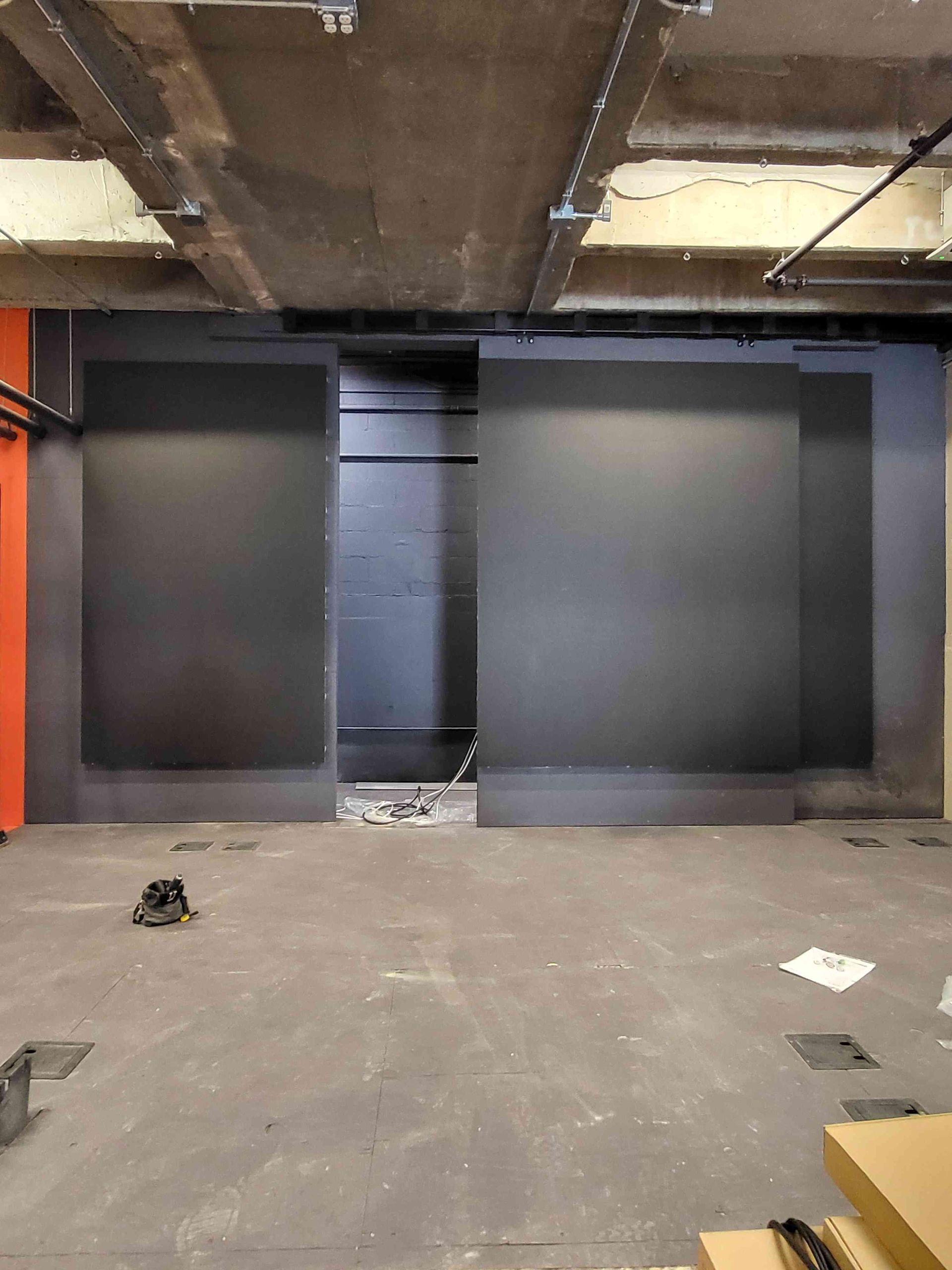 Black wall sections in an unfinished industrial space; orange wall visible on the left; gray floor and ceiling with exposed beams.