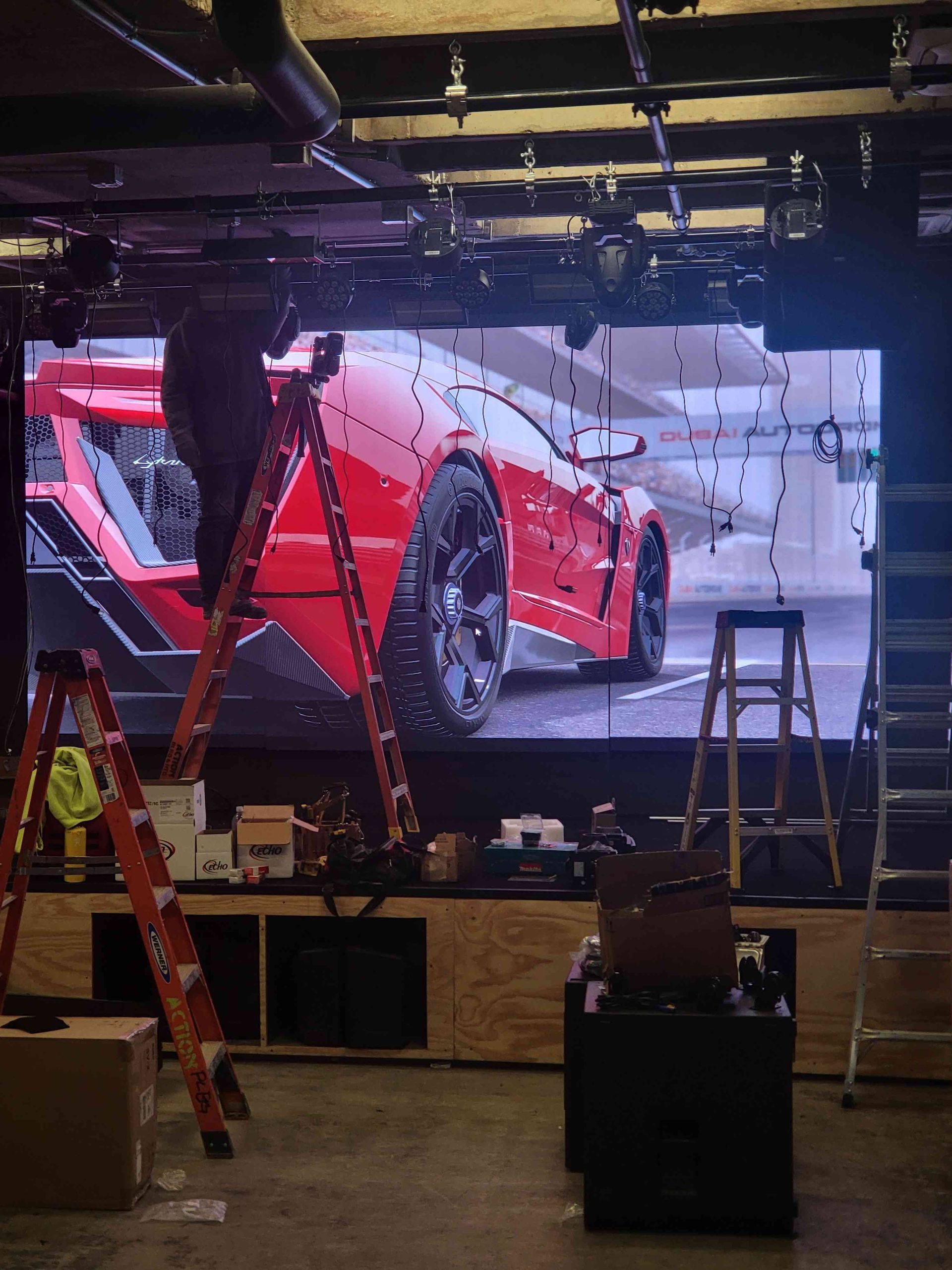 Person on a ladder adjusts a large LED screen displaying a red sports car. Inside a room with various equipment.