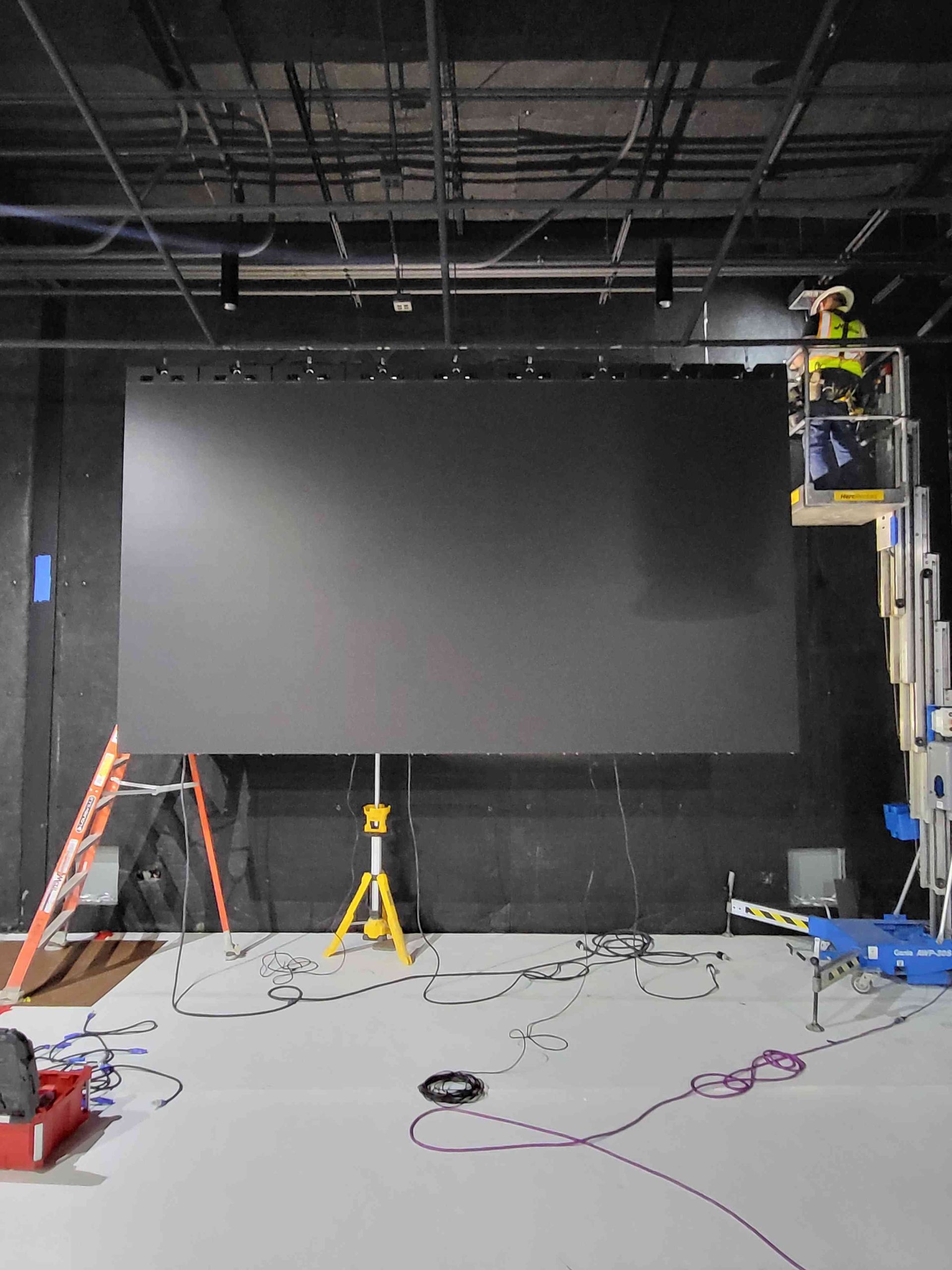 A large, dark LED screen is set up in a studio. A worker on a lift is visible, cables on the floor.