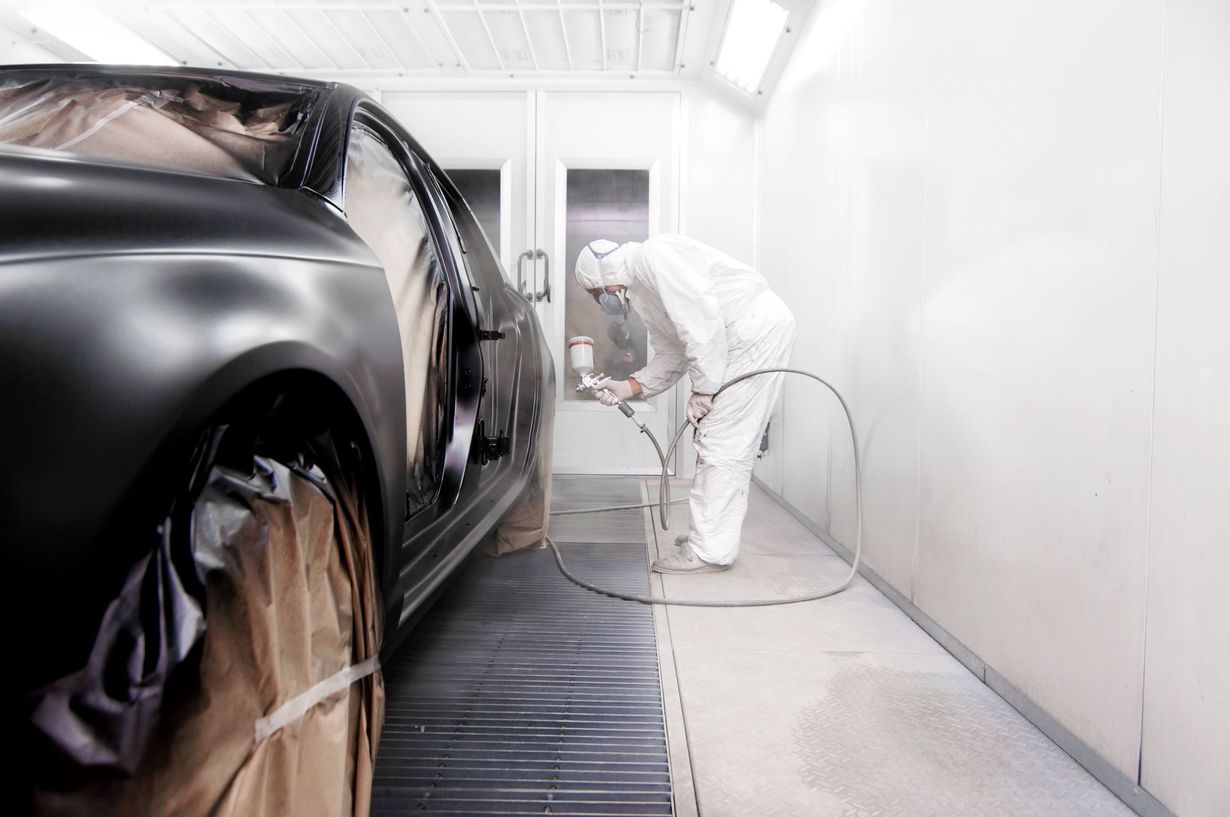 Person in protective suit spraying a black car in a white paint booth.