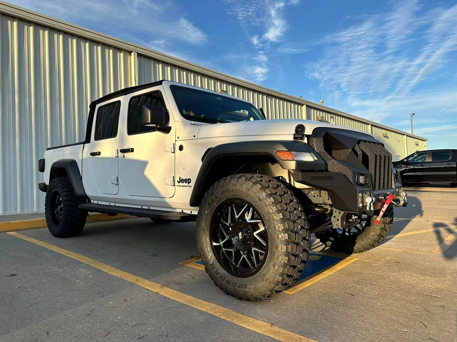 White Jeep Gladiator truck with black wheels and a custom front bumper parked in front of a metal building.