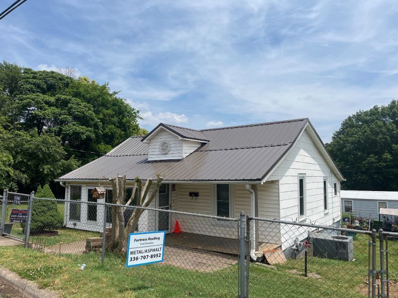 A white house with a brown metal roof is behind a fence.