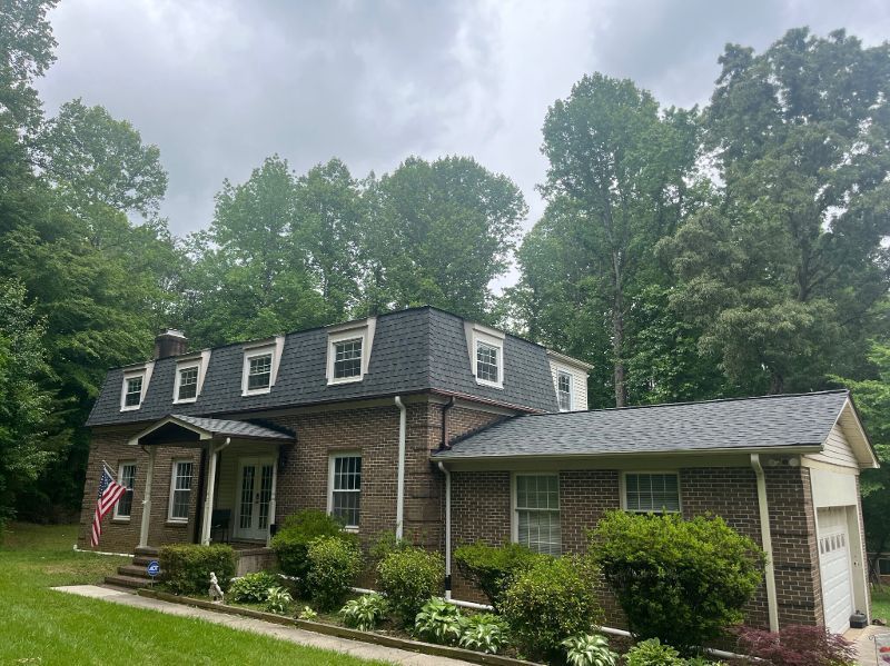 A brick house with a black roof is surrounded by trees on a cloudy day.