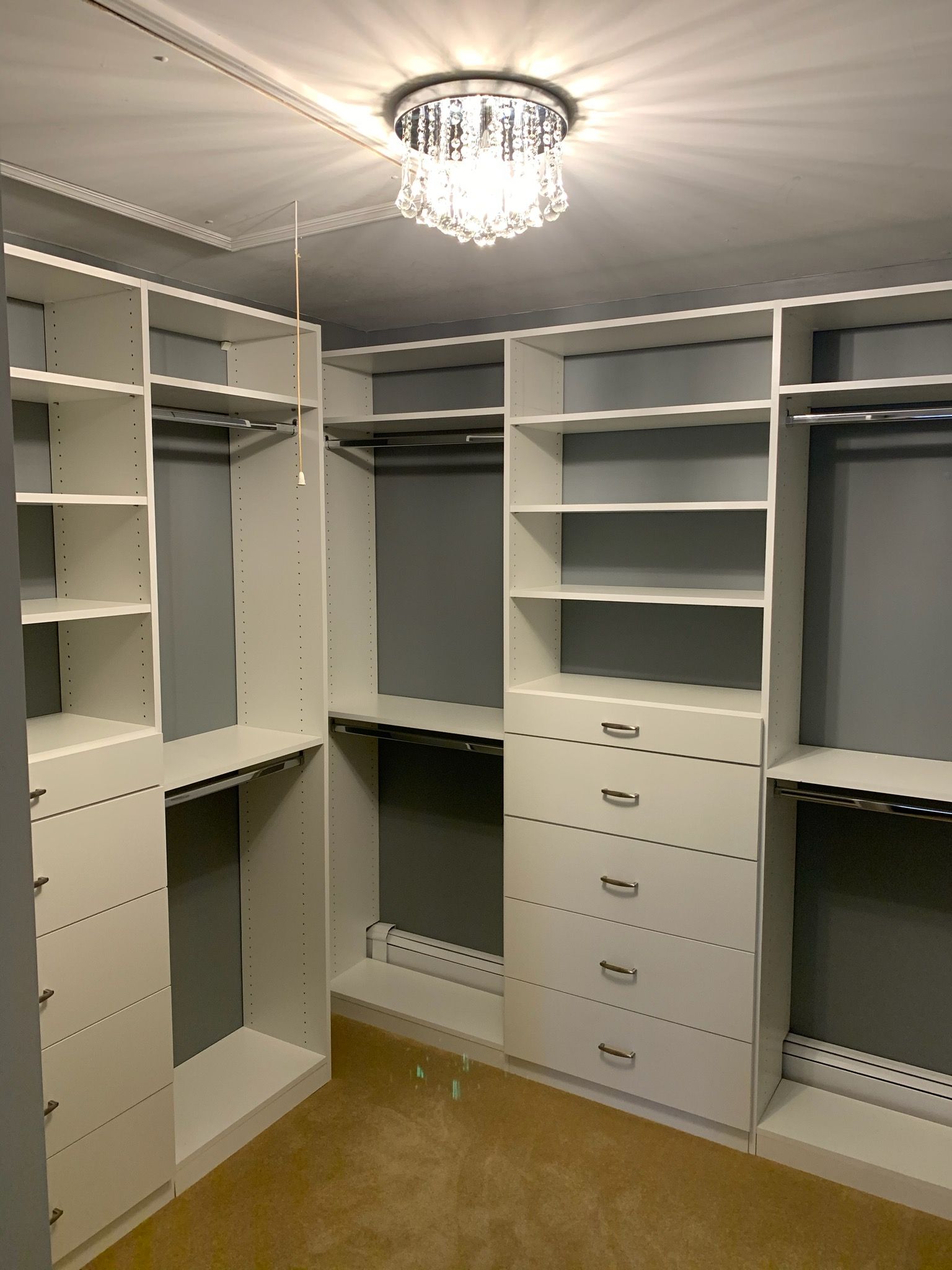 White custom closet with shelves, drawers, and hanging rods. Gray walls, beige carpet, crystal ceiling light.