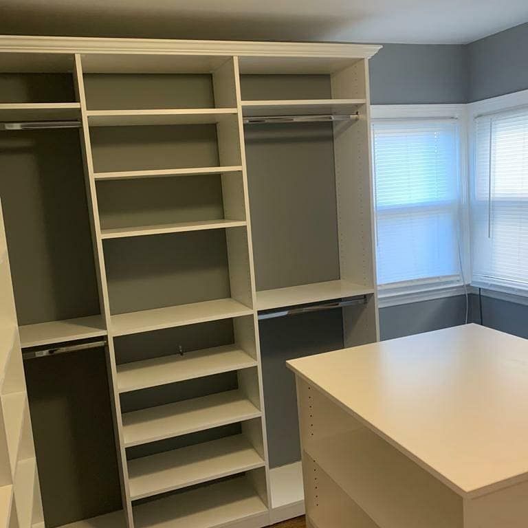 White custom closet system with shelves, hanging rods, and an island unit in a room with gray walls and a window.