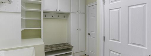 A white mudroom with cabinets, shelving, bench, coat hooks, and a closed door.