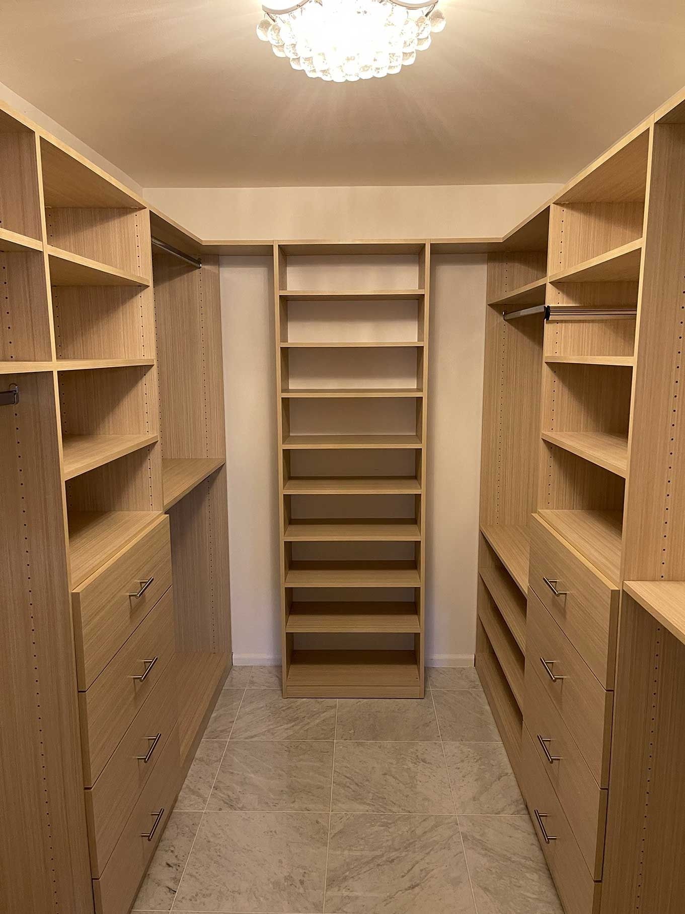 Walk-in closet with built-in light wood shelves, drawers, and hanging rods. Central vertical shelving unit and neutral tile flooring.