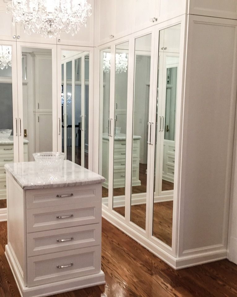 White walk-in closet with mirrored doors and marble-topped island on wood floor.