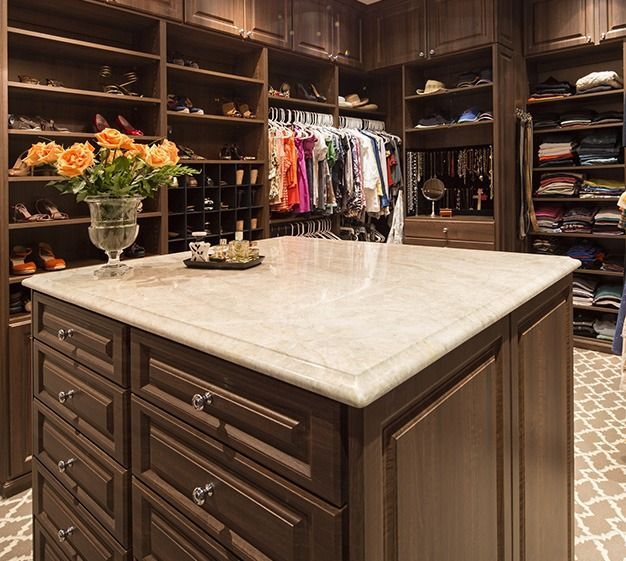 Walk-in closet with island. Brown cabinetry, beige countertop, shoes on shelves, hanging clothes, bouquet.