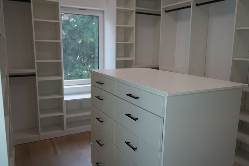White walk-in closet with built-in shelves and drawers. A window is in the center, and black handles are on the drawers.
