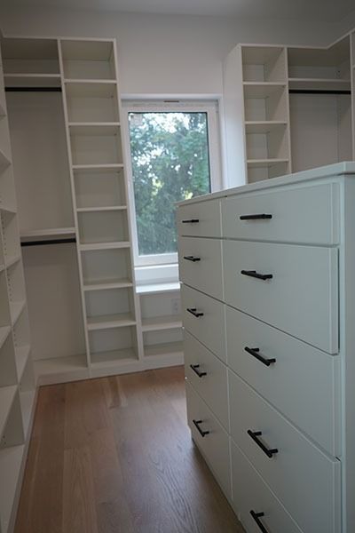 White walk-in closet with shelves, drawers, and window. Wooden floor, black drawer pulls, and hanging rods.