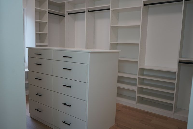 White built-in closet with drawers and shelves. Black handles, hanging rods, and light-colored wooden floors.