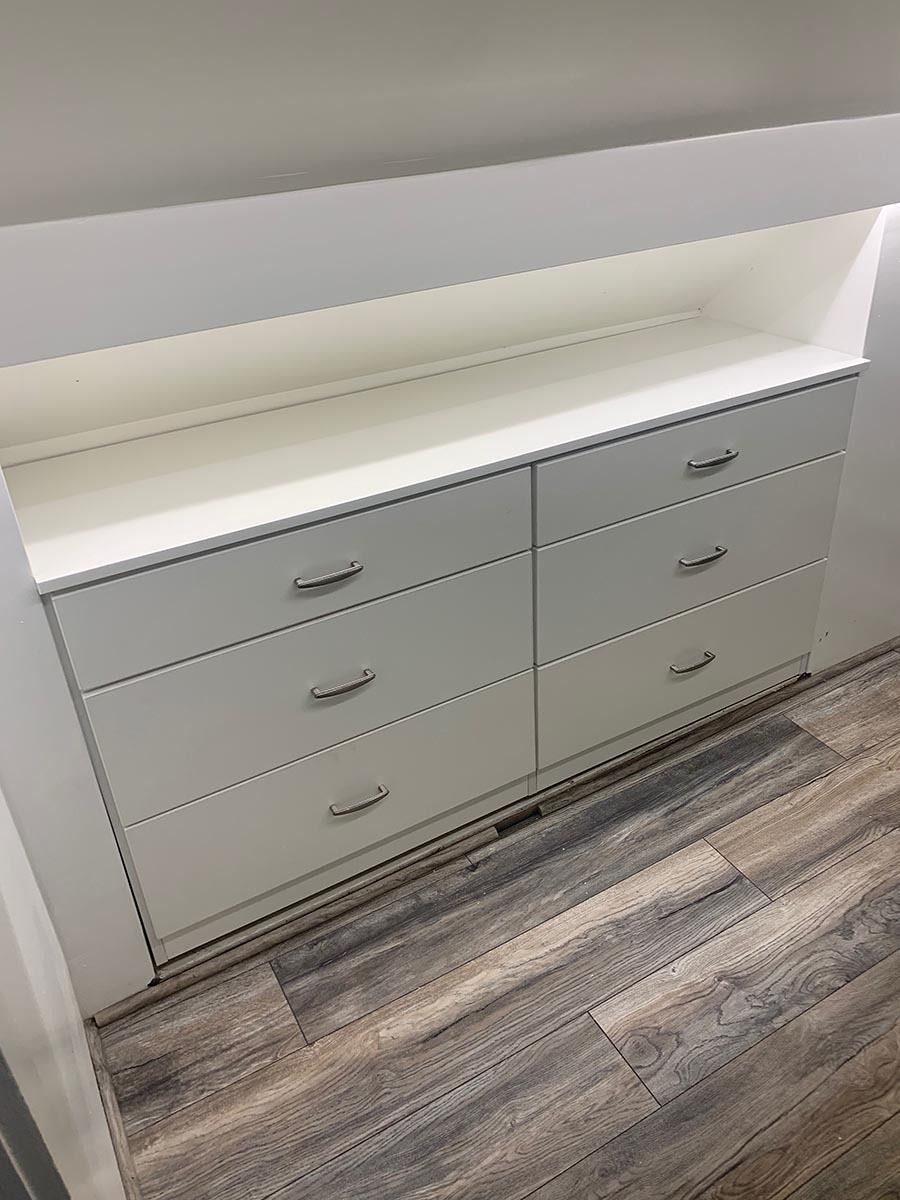 White dresser with six drawers, silver handles, built into an alcove with a shelf above.