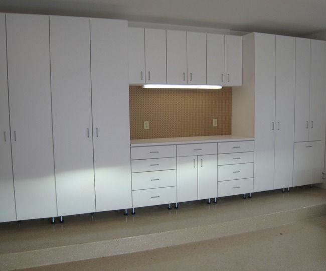 White storage cabinets and workbench in a garage with tan walls, containing a pegboard and overhead lighting.