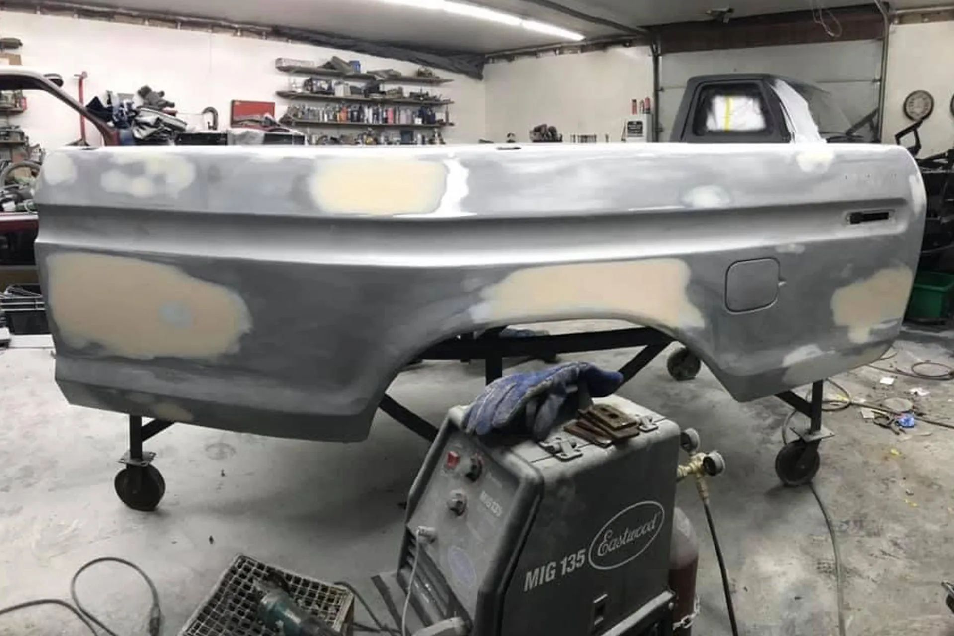 Truck bed in a garage, undergoing bodywork with filler patches. Gray, metal, and tan colors.