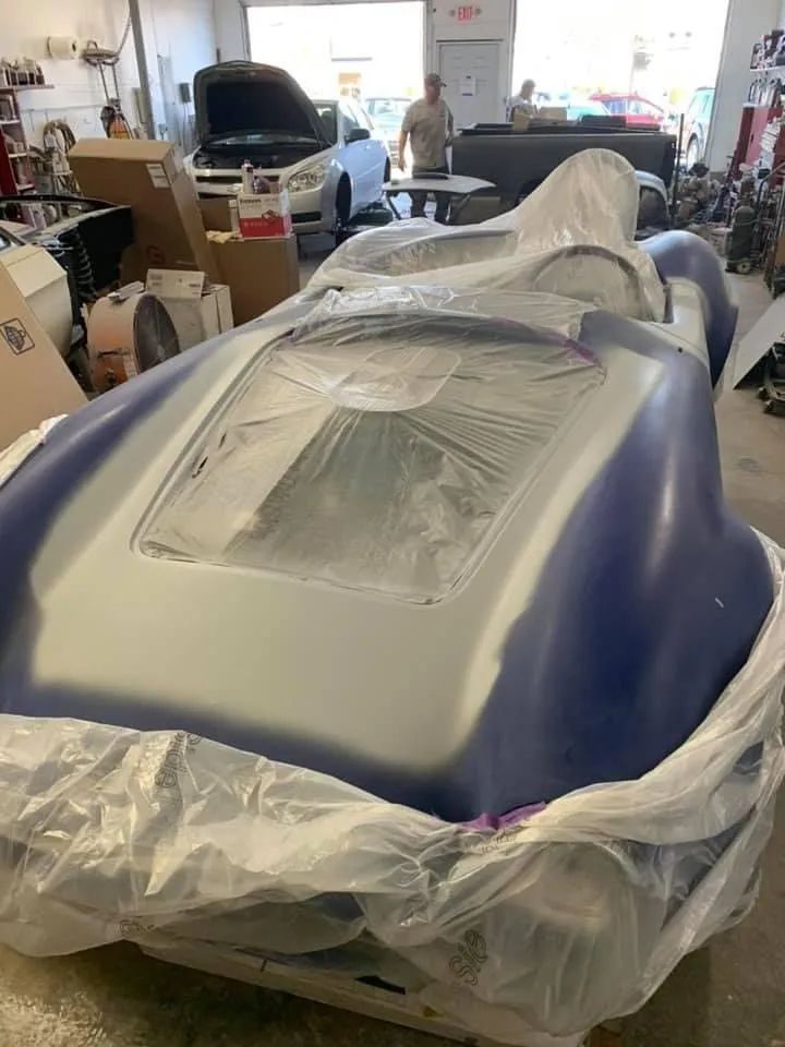 Blue and silver car body wrapped in plastic inside a workshop, a person in the background.
