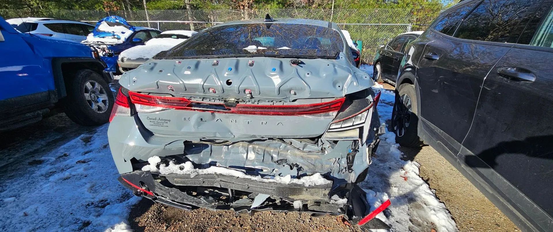 Rear end of a severely damaged grey sedan in a lot, surrounded by other vehicles.