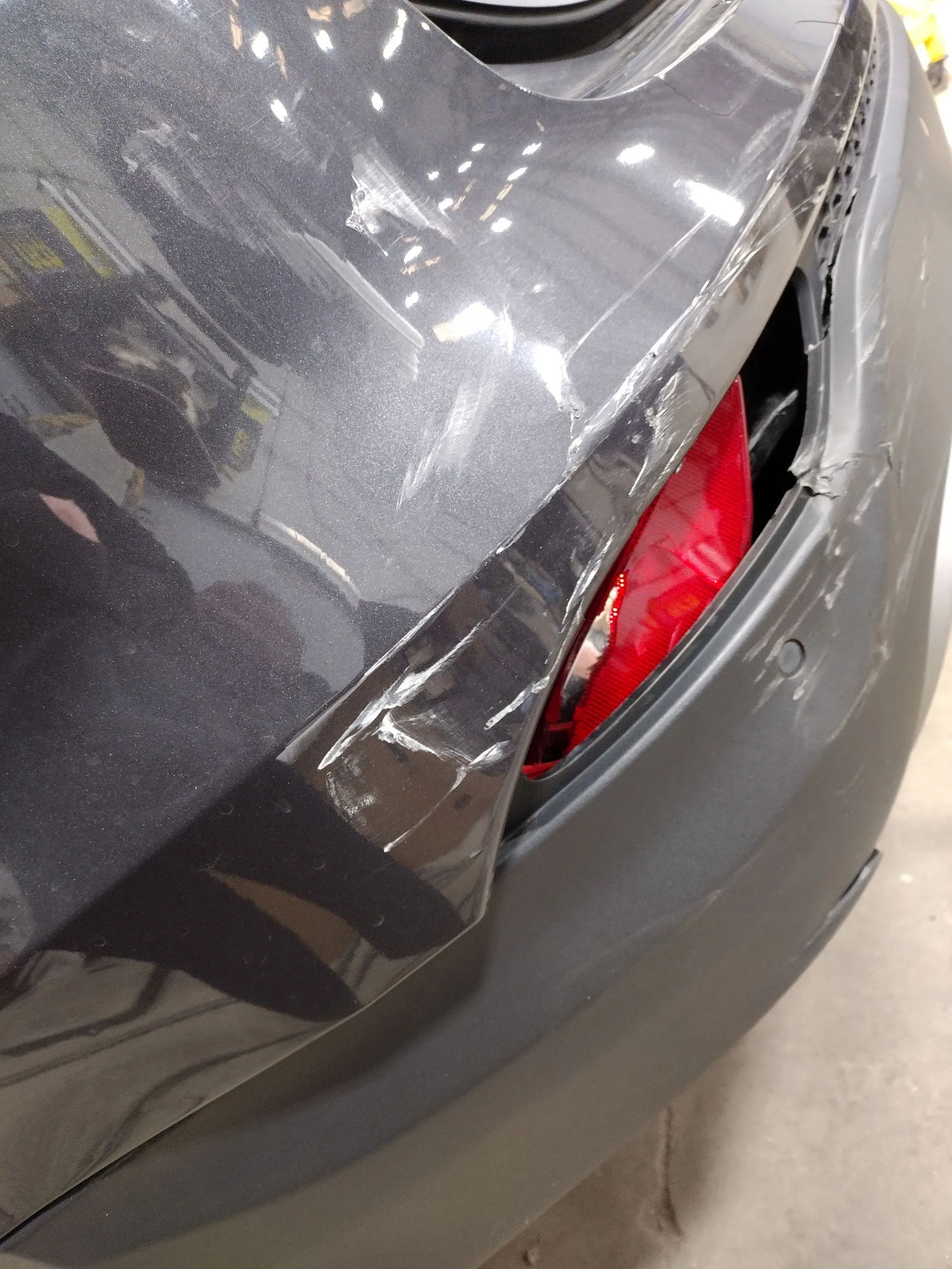 Damaged gray car bumper. Tail light exposed, with white scrapes and dents on the side panel.