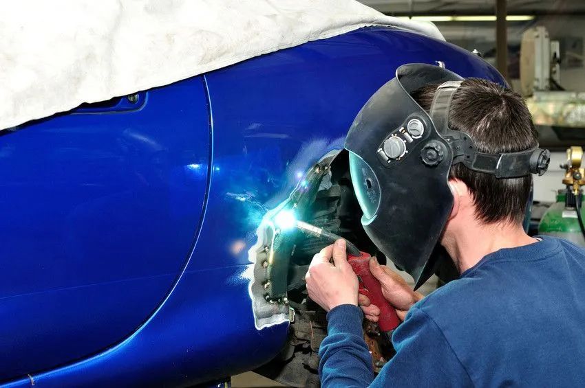 A person welds a blue car panel, wearing a welding mask and holding a welding torch.