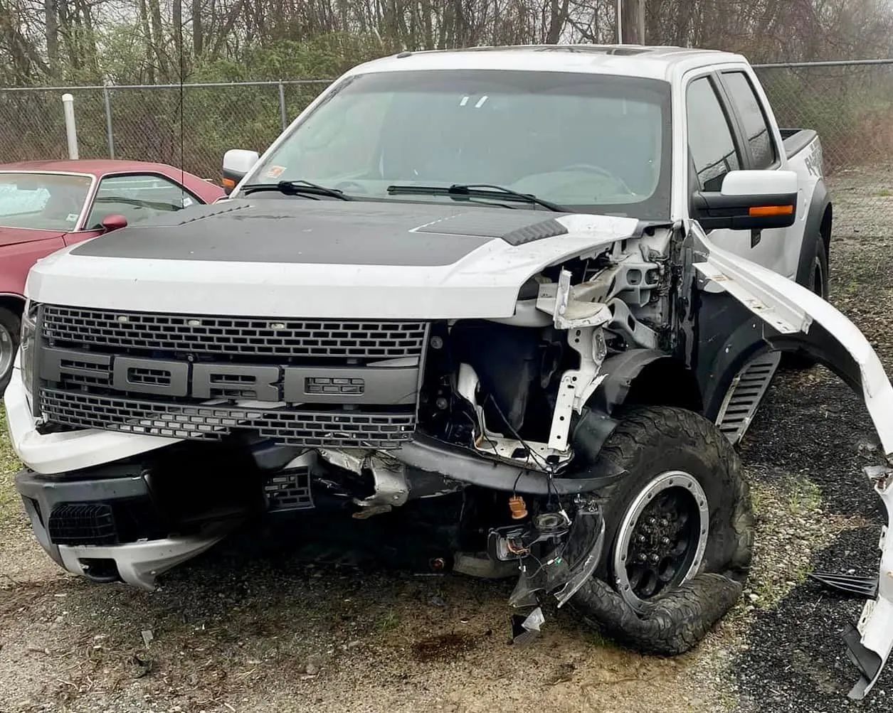 A white Ford Raptor pickup truck with significant front-end collision damage, including a flat driver-side tire.