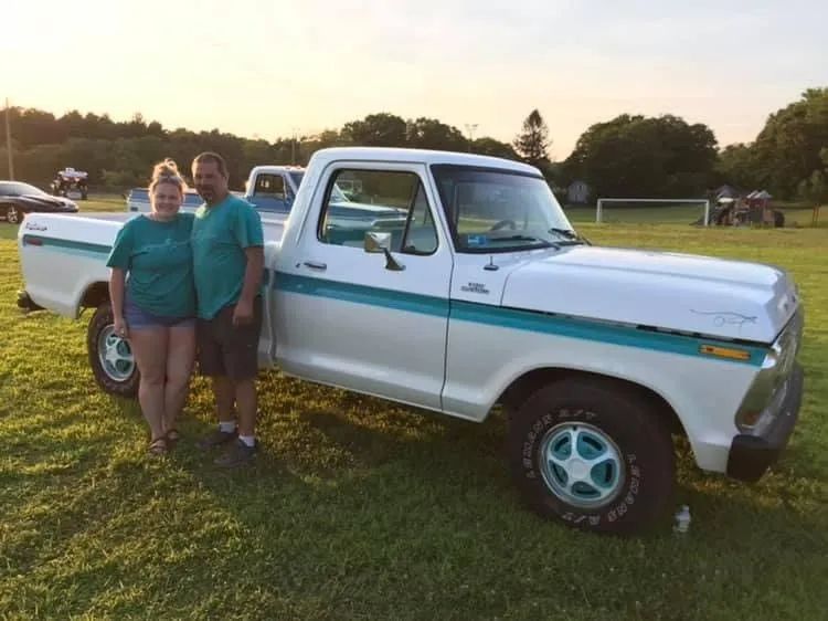 Couple poses with a white and teal Ford pickup truck on a grassy field.