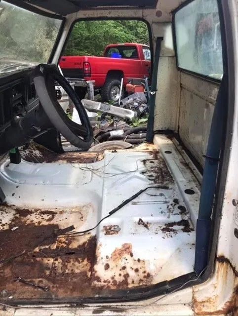 Interior of a rusty, white truck cab with a red truck visible through the open door.