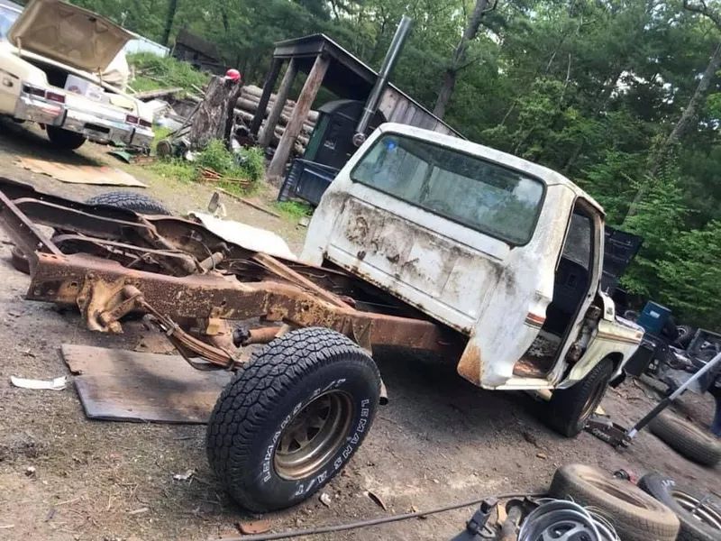 Rusty truck chassis with a white cab, tire, and parts in a yard.