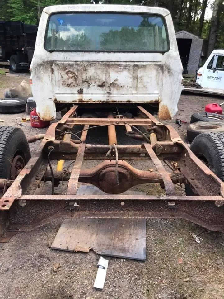 White truck cab perched on a rusty chassis, outdoors. Tires, tools, and a shed are nearby.
