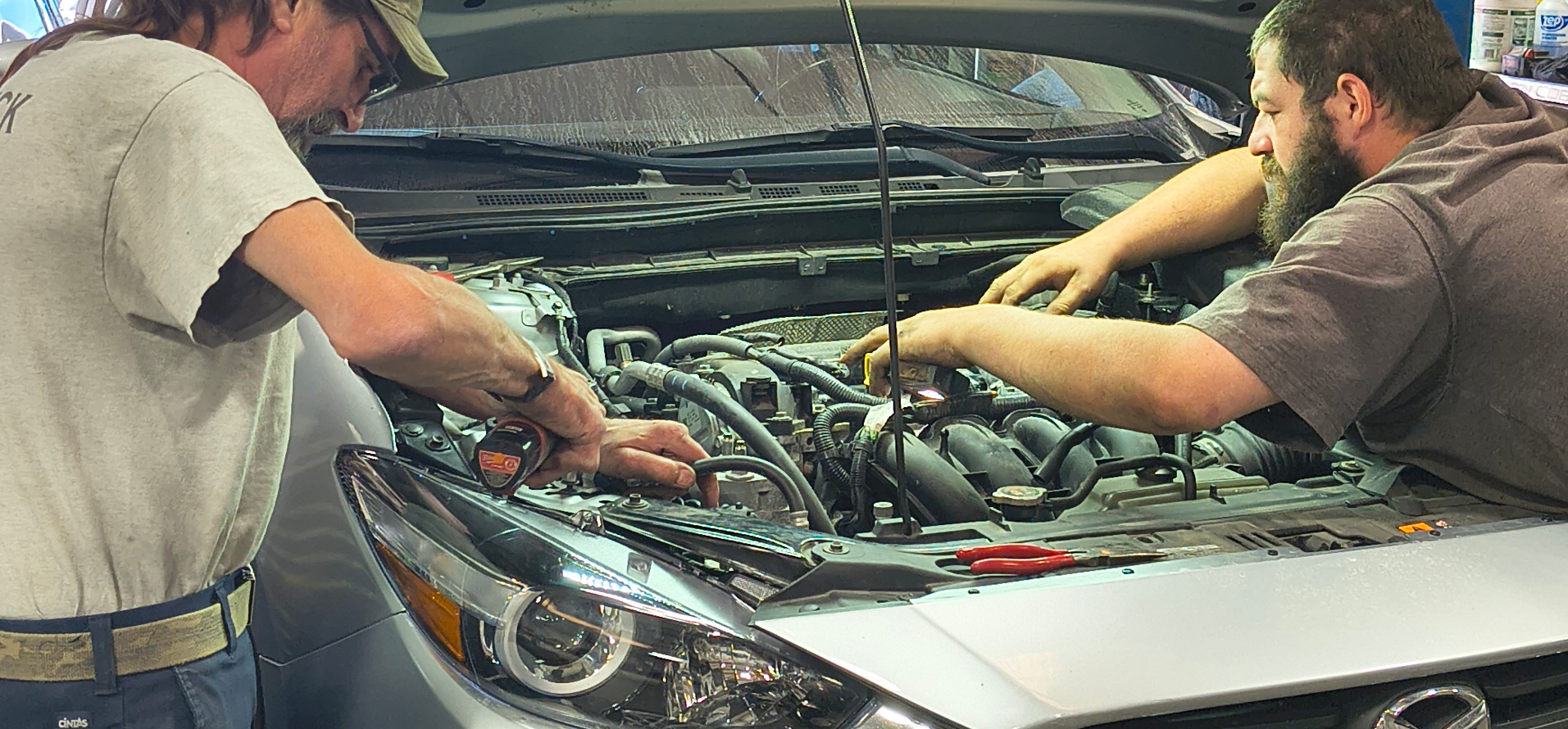 Two mechanics working on a car engine in an auto shop. One wears a baseball cap.
