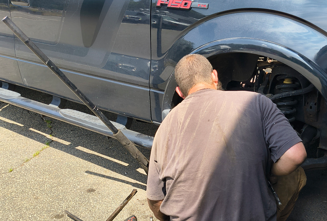Person working on the front wheel of a dark-colored vehicle, outdoors. A long lever sits on the ground.