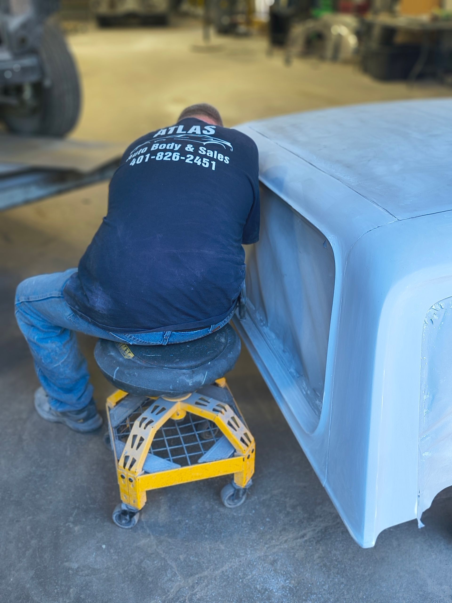 Person in a shop, working on a vehicle's body, seated on a yellow rolling stool.