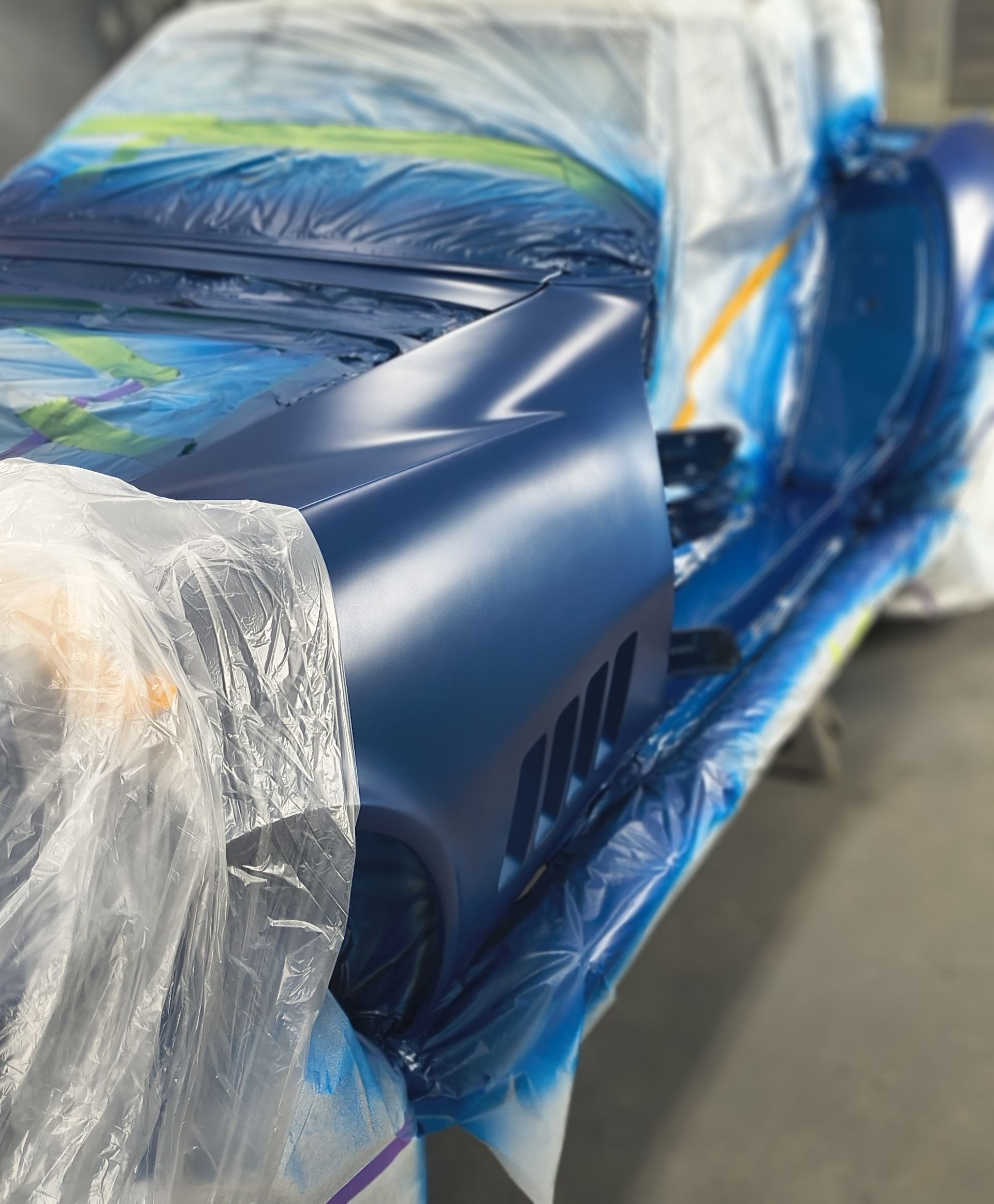 Blue car body, freshly painted with matte finish, covered with plastic sheeting in a workshop.