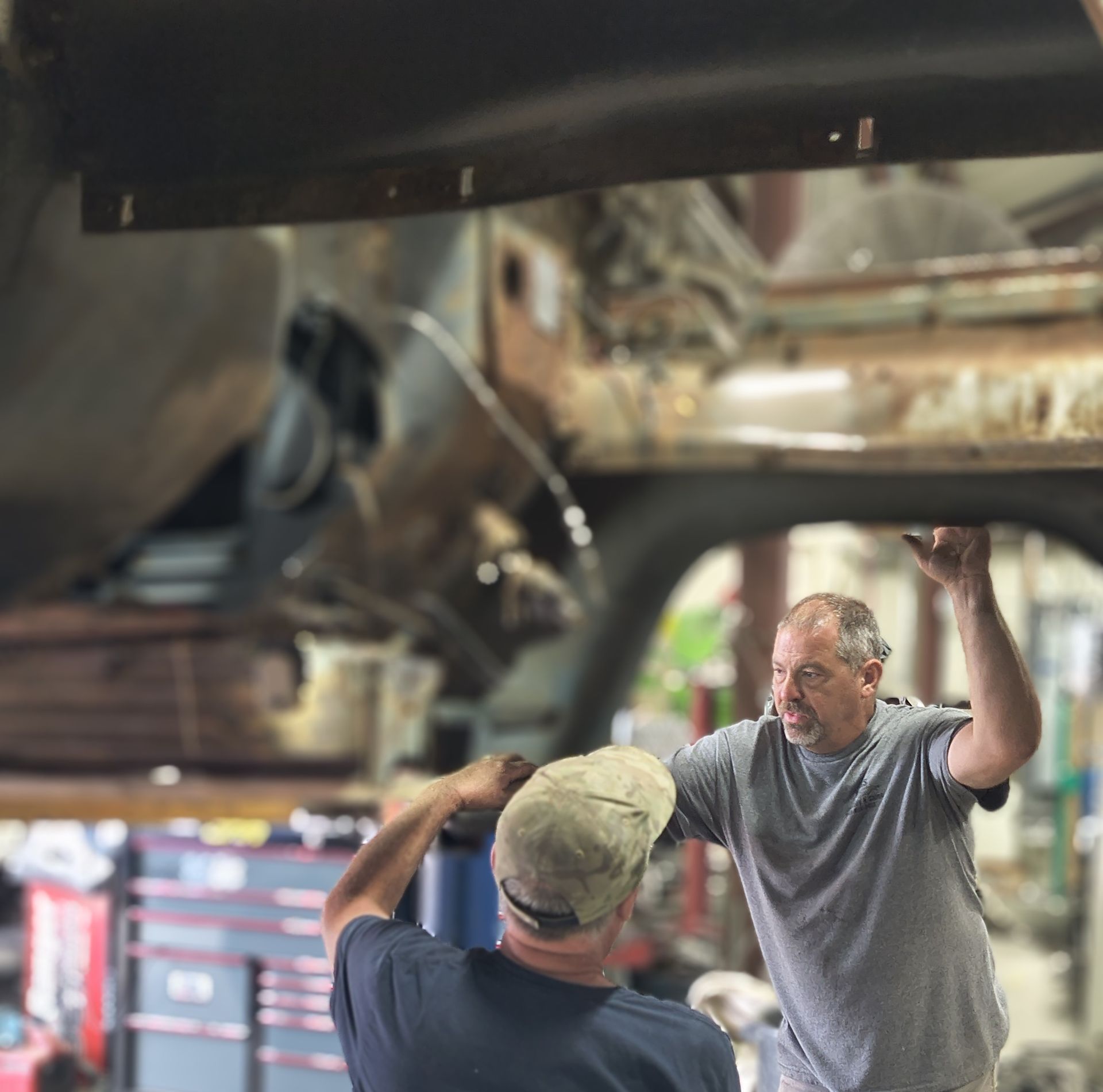 Two men working under a vehicle in a shop, one looking up, the other facing away holding something.