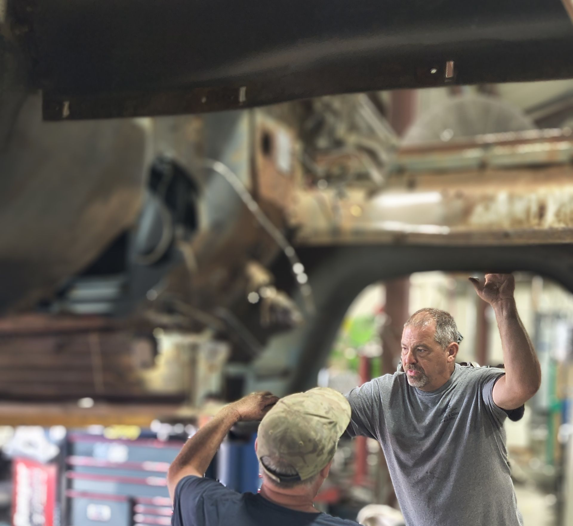 Two people stand in an auto shop, inspecting the underside of a car chassis suspended on a lift.