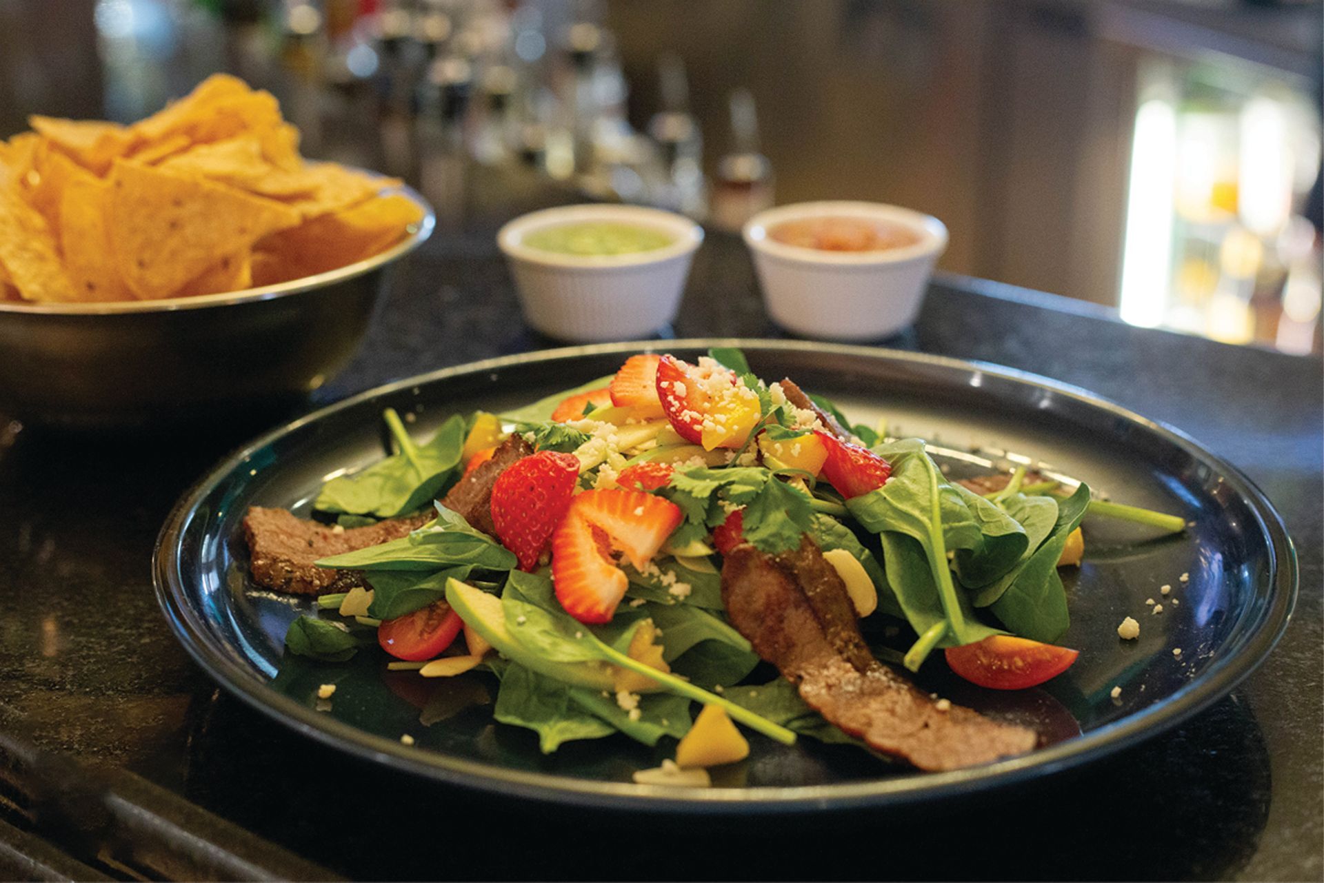 a black plate topped with a salad and strawberries next to a bowl of tortilla chips