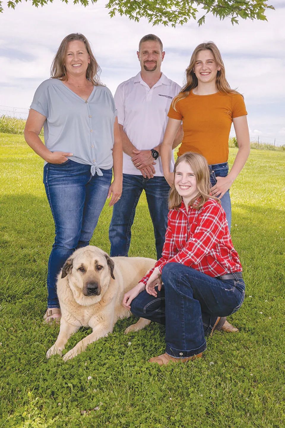 Family of four and a dog pose in a grassy field. The two women kneel and stand, and the man and dog are also present.