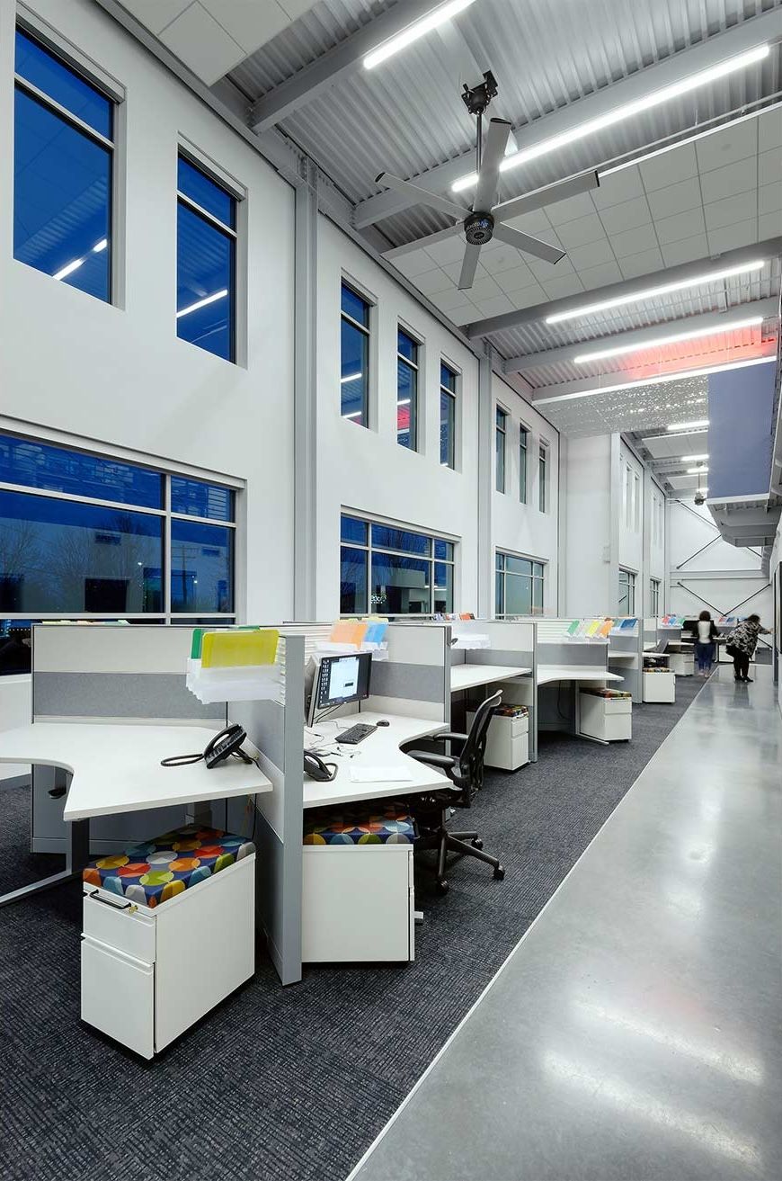 Modern office space with rows of cubicles, tall windows, and a ceiling fan.