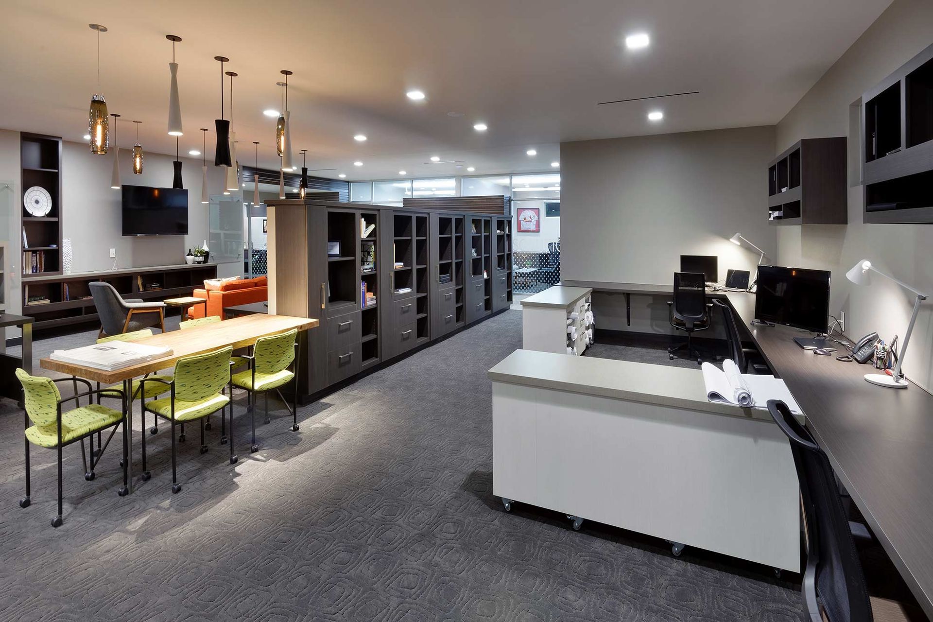 Office space with desks, shelves, chairs, and lighting. Contains tables with green chairs, and monitors.