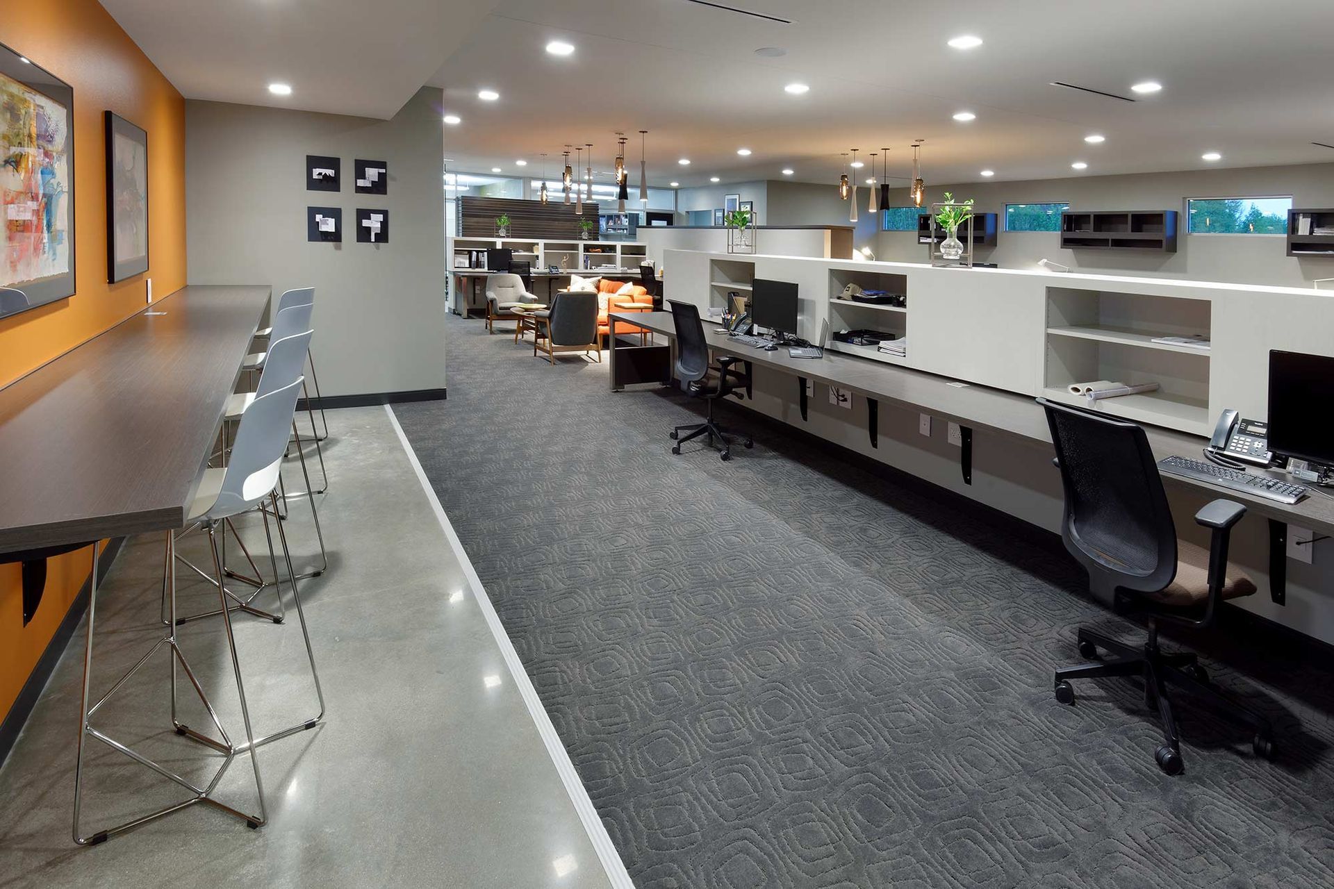 Modern office with desks, computers, chairs, and a long counter with stools. Gray carpet, orange and gray walls.