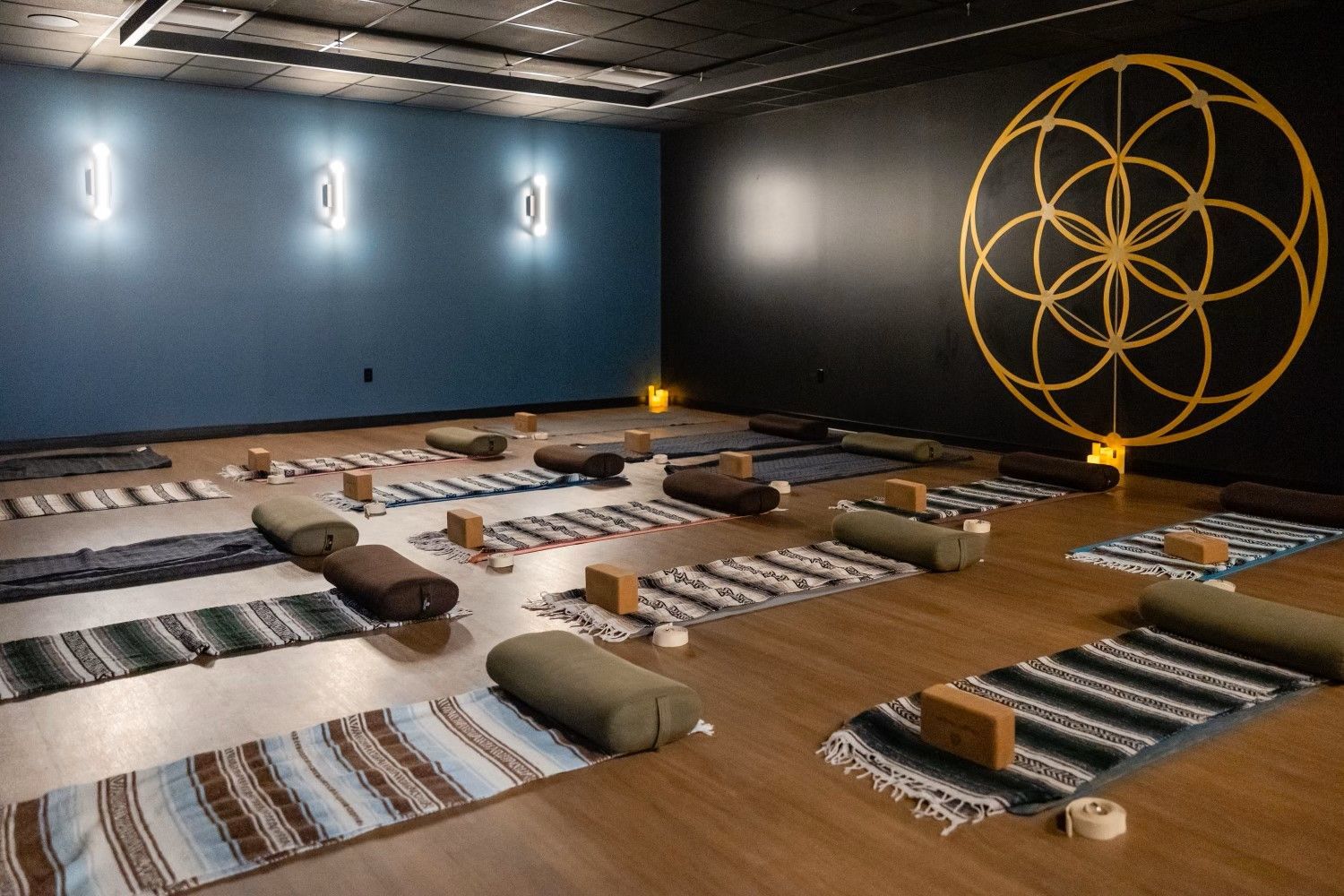 Yoga studio: mats, bolsters, and blocks laid out on wood floor. Black and blue walls with a gold geometric symbol.