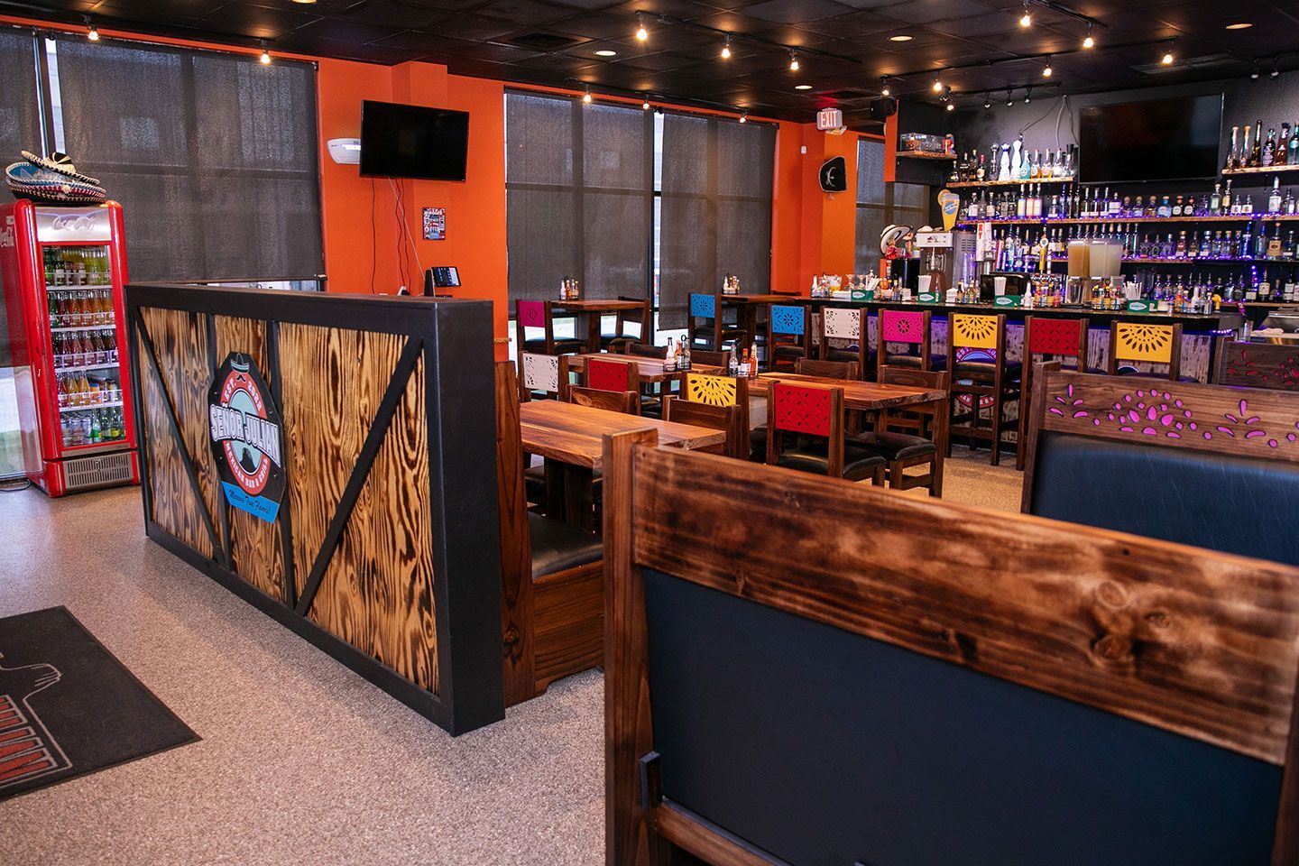 Inside a bar with wooden booths, colorful chairs, and a fully stocked bar. Orange walls and a drink cooler are also present.