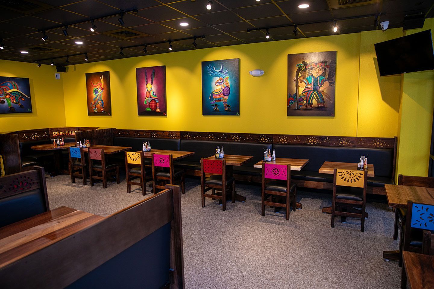 Restaurant interior with yellow walls, colorful art, and wooden tables with chairs.