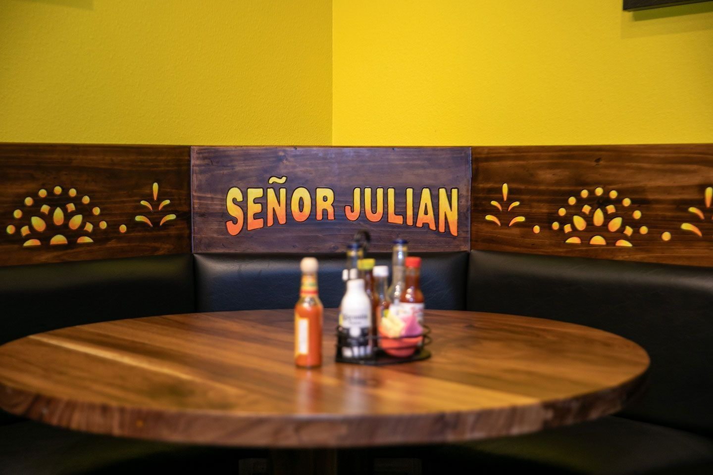 Round wooden table in a booth at Señor Julian restaurant, with bottles of hot sauce; yellow wall, wooden sign.