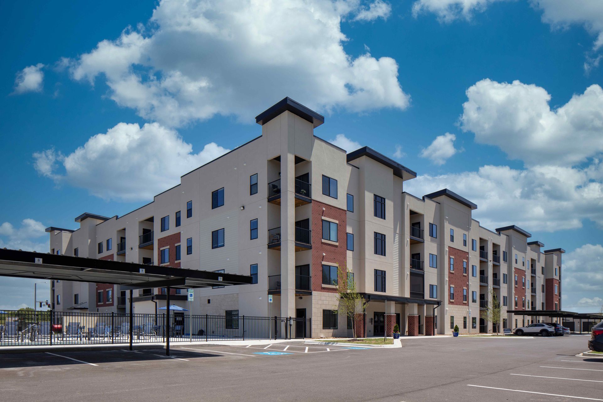 Multi-story apartment building on a sunny day with blue sky and puffy clouds, parking in foreground.