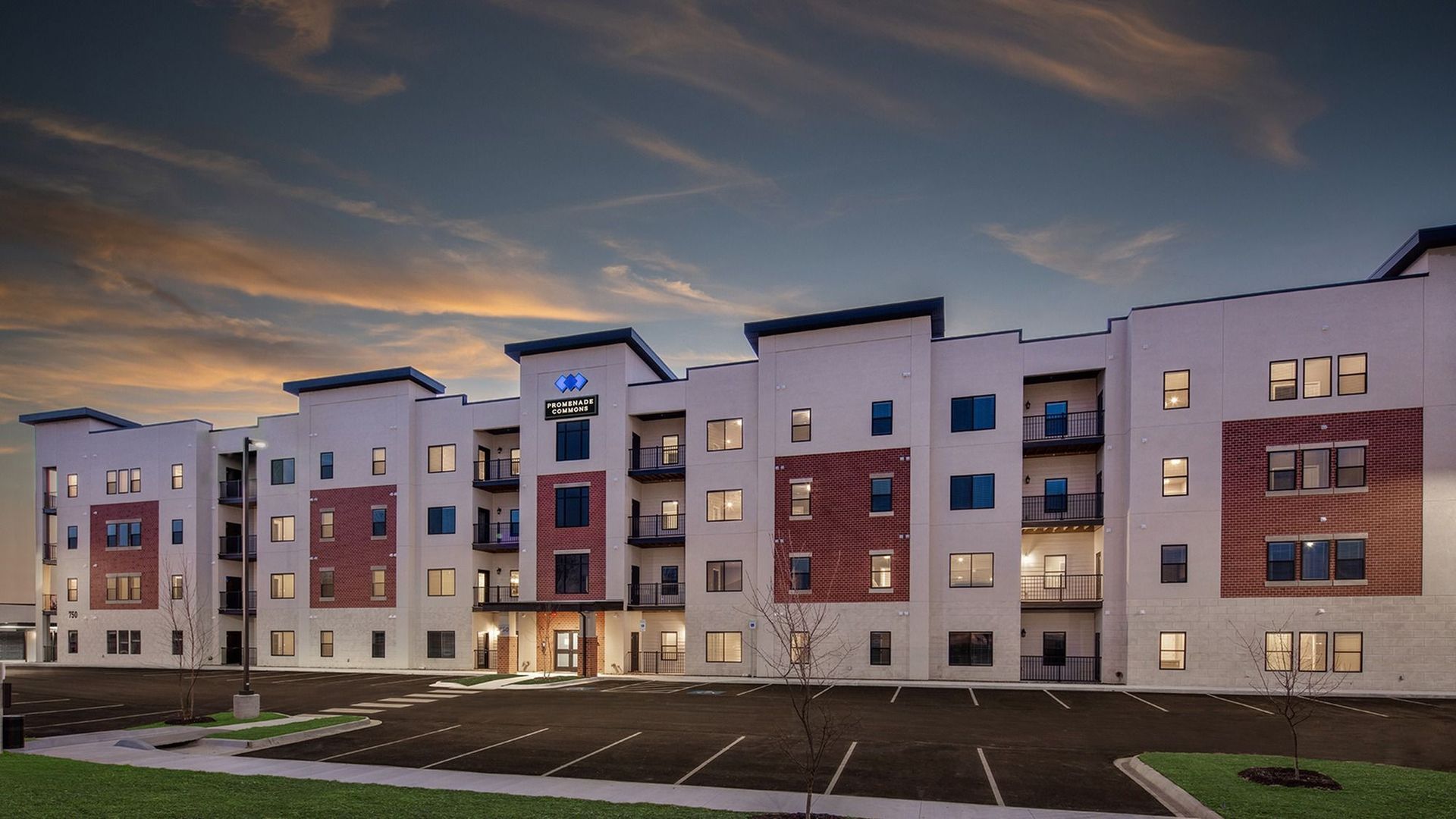 Multi-story apartment building with brick and white exterior, parking lot, and sunset sky.